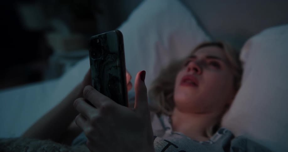 Blonde woman lying in bed uses smartphone crying bitterly in bedroom at night