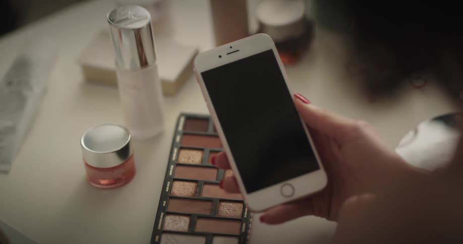 White woman sitting at a table with a makeup palette and comestics on top of it tapping on her smartphone