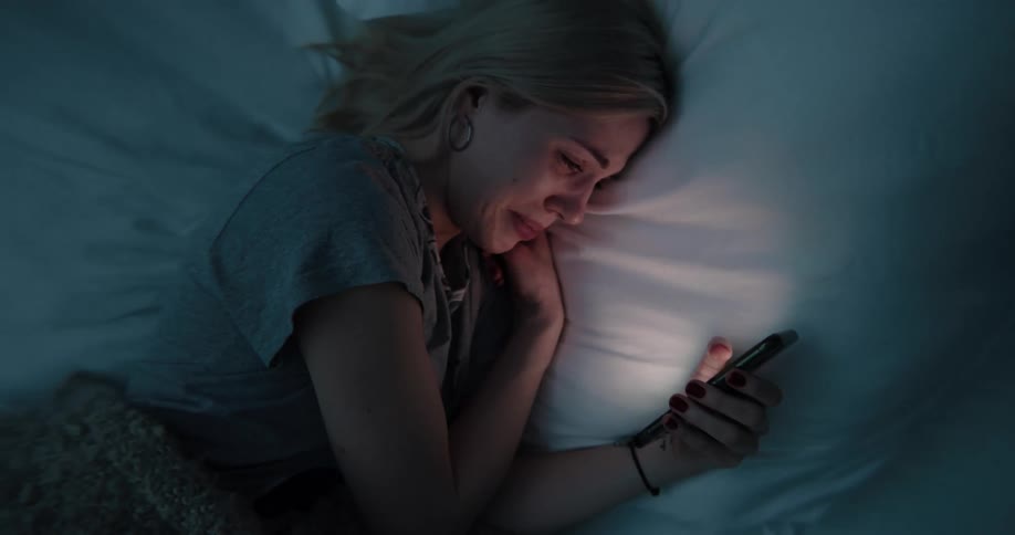 A young woman cries while scrolling through her phone in bed in a dark bedroom