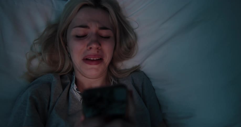A teenage girl lying in bed takes her phone out from under her pillow and looks at it and cries and puts it back under the pillow and rolls up in to a ball