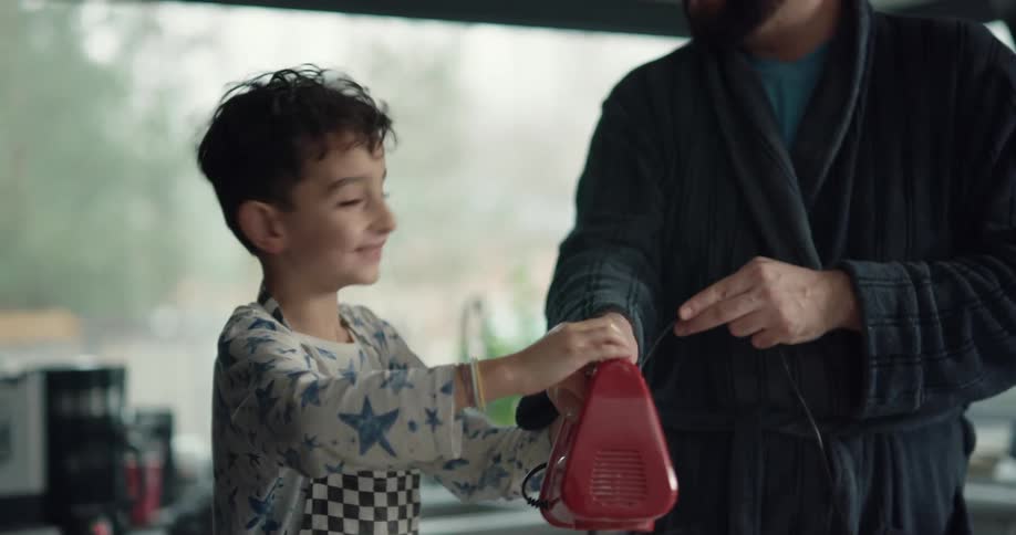 A boy holds a handheld mixer together with his father while mixing batter in a home kitchen during the day