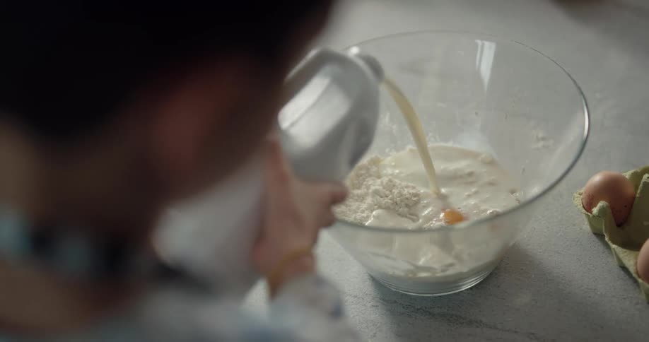 Overhead view of a boy pouring milk on flour and eggs in a glass bowl