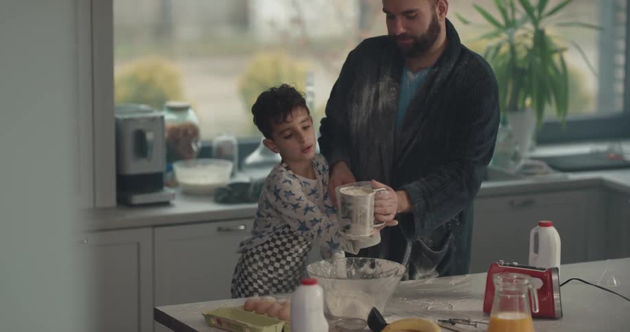 A dad helps his boy sift flour for a batter in the kitchen
