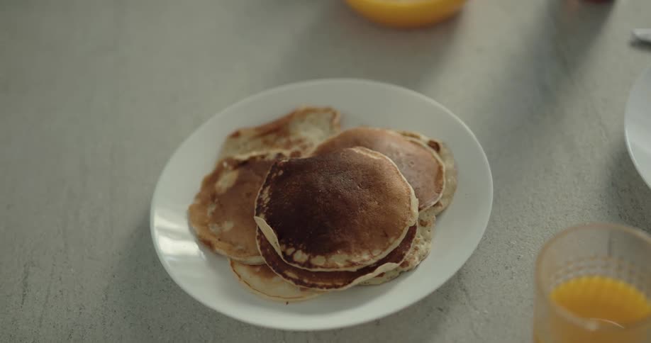 A person using a knife to take a pancake from a white plate in a table with a orange juice cup next to it