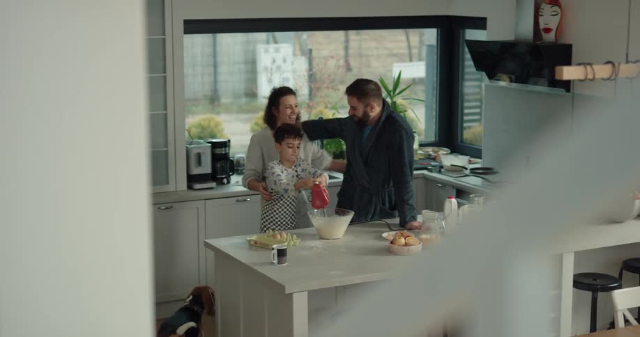 A young boy stands by his dad in the kitchen and mixes batter when his mom walks downstairs drinking coffee and gives them both kisses