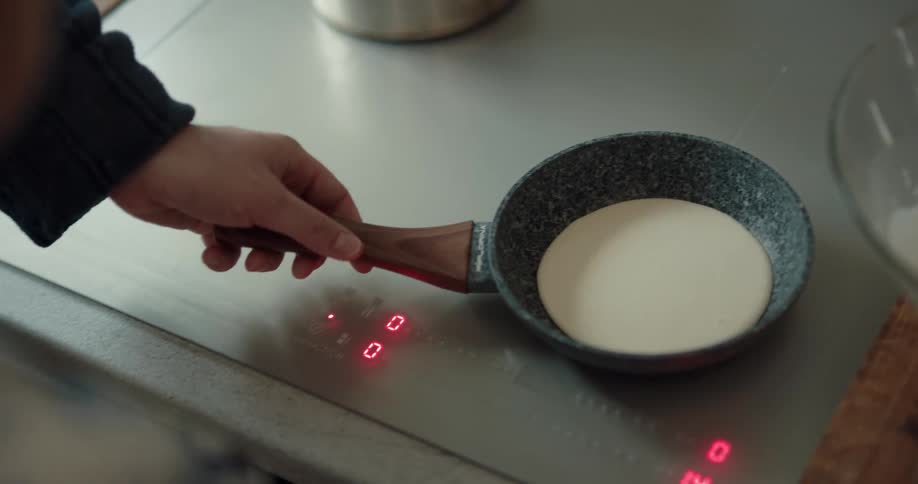 A man holds a pan on a cooktop as his son pours pancake batter in it