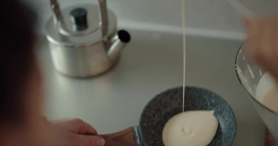 A man holds a pan on a cooktop while his daughter puts pancake batter in the pan