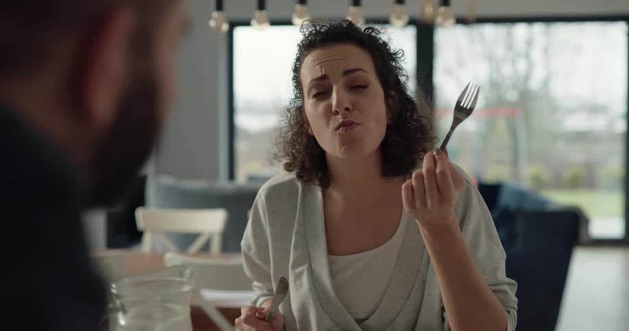 White woman next to a kitchen counter tasting some food and enjoying it while talking to a man