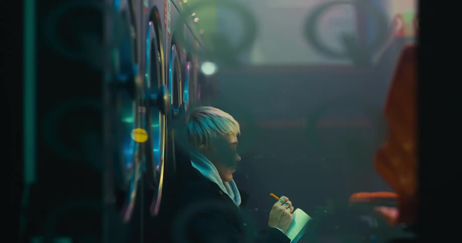 A woman writes in a notebook while sitting against a laundry machine in a laundromat at night
