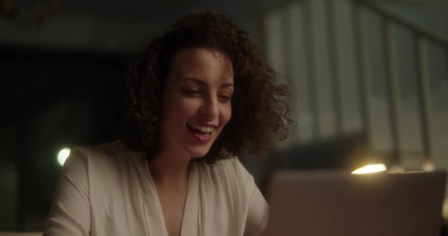 Woman with curly hair and white blouse sits at table using laptop laughing and hiding her face at night
