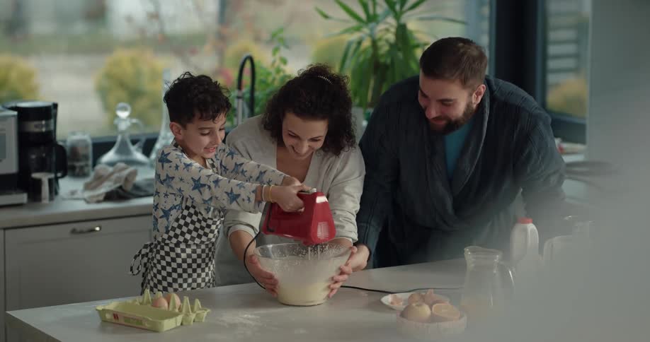 A boy uses a mixer to stir batter in a mixing bowl being held in place by his mother as his father watches from beside her