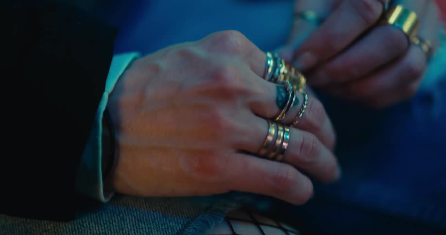 A man is sitting indoors and fidgeting with his rings and fishnet stockings