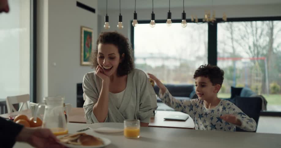 Young boy leads his mother to the kitchen table and after she sits down her husband places a plate of pancakes on the table