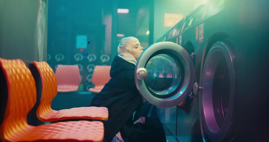 Young blonde woman enters a laundromat and starts filling a washing machine with laundry