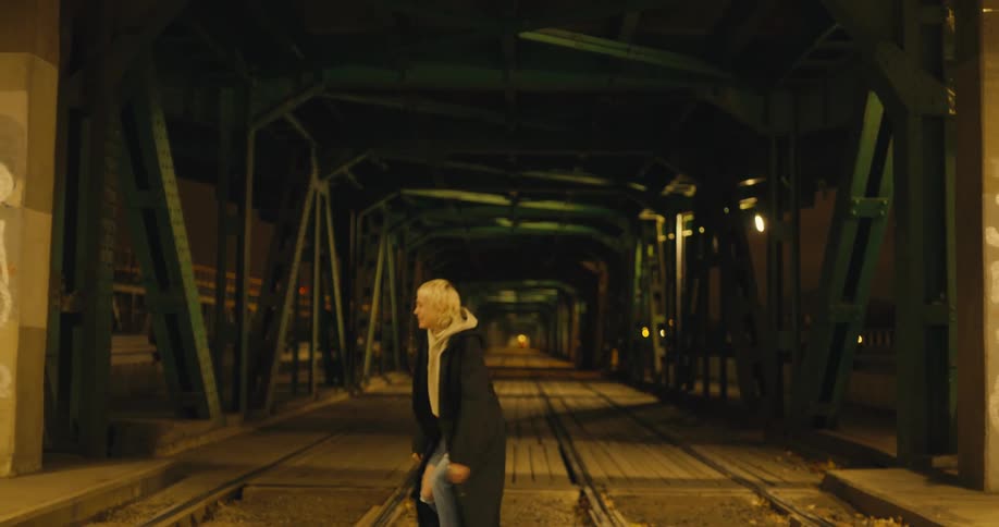 Smiling blonde woman wearing long coat walks out of train tunnel and sits between tracks