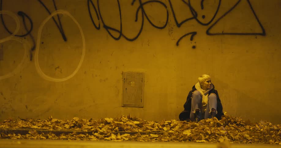 Young blonde woman sitting over a pile of dry leafs reclined against a wall near a street