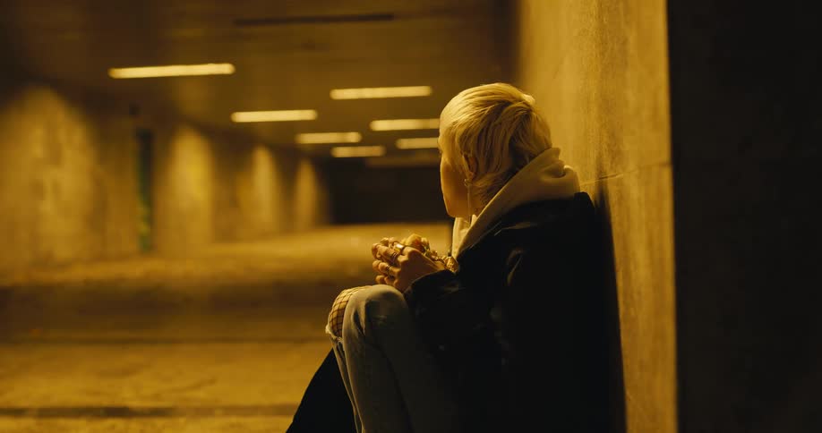Blond teenaged girl sits against the wall of a tunnel and eats a sandwich