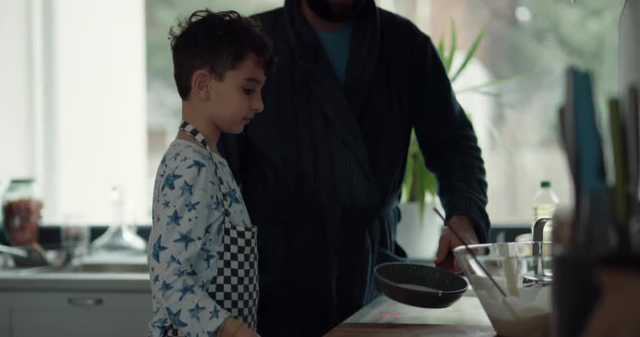 A boy jumps up on a step stool to ladle batter into a pan helping his dad make breakfast