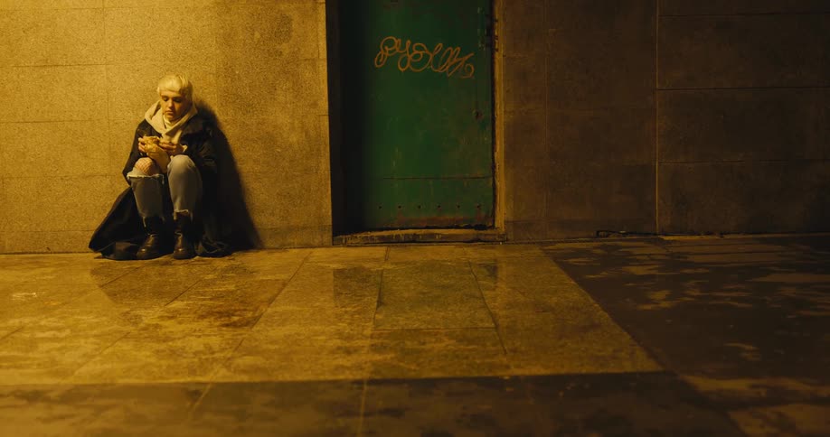 A woman sits down against a building wall and opens a paper bag to take a bite of food