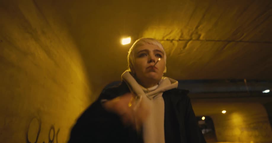 Young short haired woman lights a cigarette walking under a bridge in the city at night