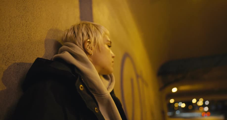 A young woman lights and smokes a cigarette while leaning against the wall under an overpass in a city at night