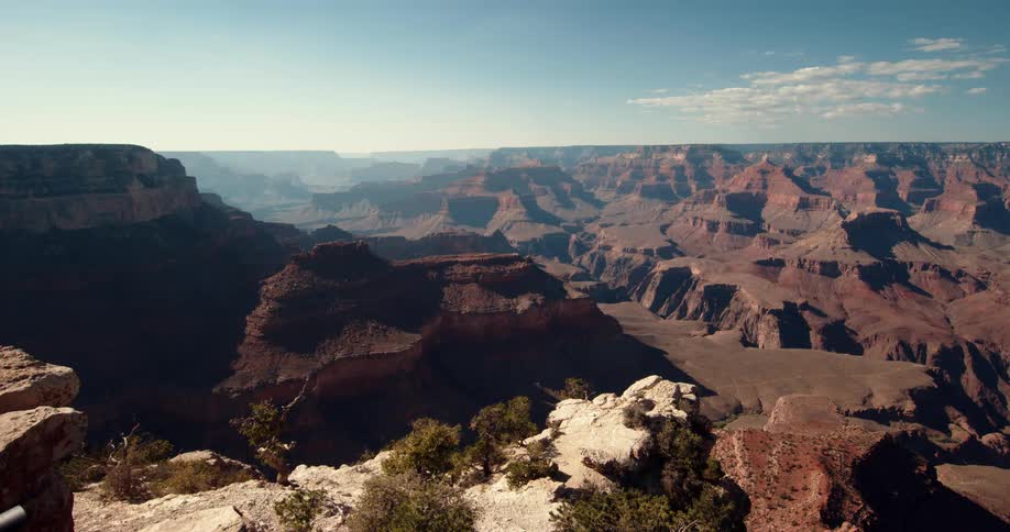 The sun shines on trees high on a mountain ledge and there are mountains and rock formations in the distance