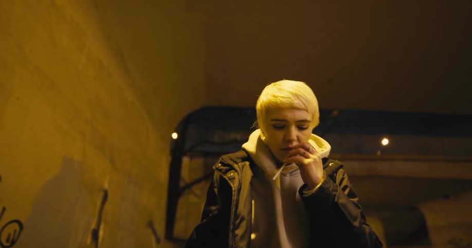 A teenage girl closes her coat around her as she walks under a bridge with a train above her and then lights a cigarette as she continues walking