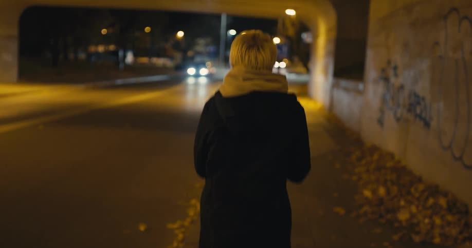 Blonde woman rushes along sidewalk under bridge and keeps looking back