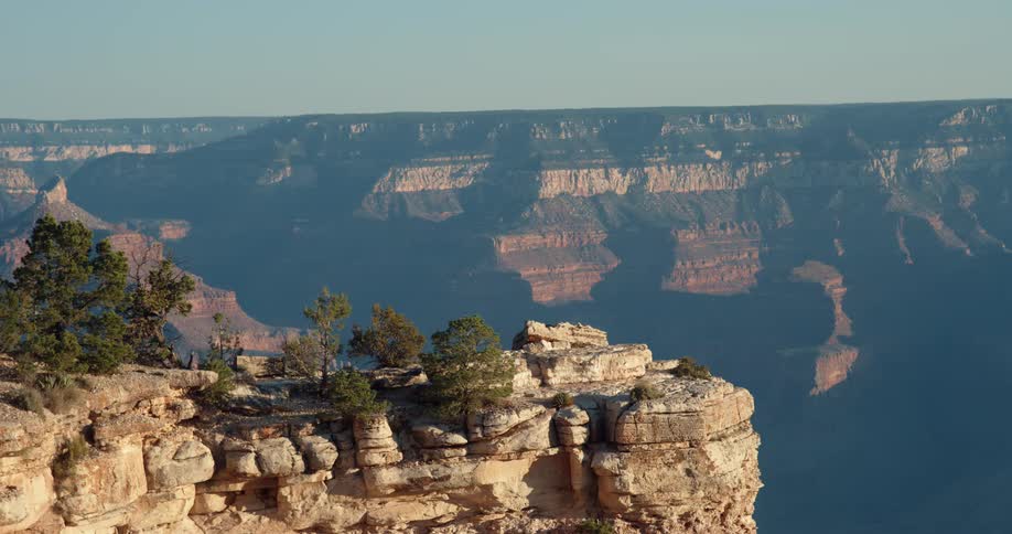 The sun shines on trees high on a mountain canyon and there are mountains and rock formations in the distance under a clear sky