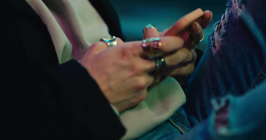 Young blonde woman sitting near a row of red seats on a laundromat at night texting on her phone