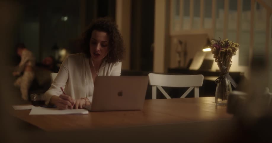 White woman sitting at the table on a video call on her laptop bothered by her husband and son that are playing behind her on the couch
