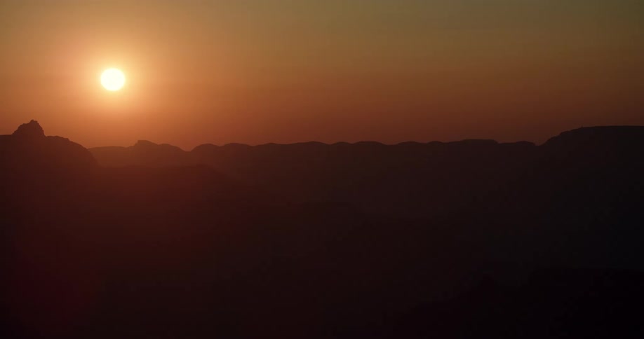 The sun shines brightly above silhouettes of mountains in an orange sky