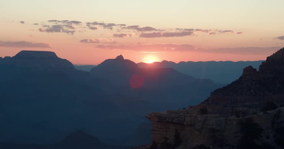 A bright sun is on the horizon behind silhouettes of mountains in a hazy sky