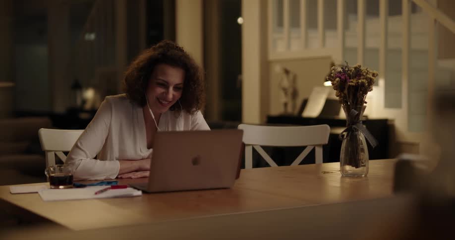 White woman sitting at the table inside her house wearing headphones and doing a work related video call on her laptop