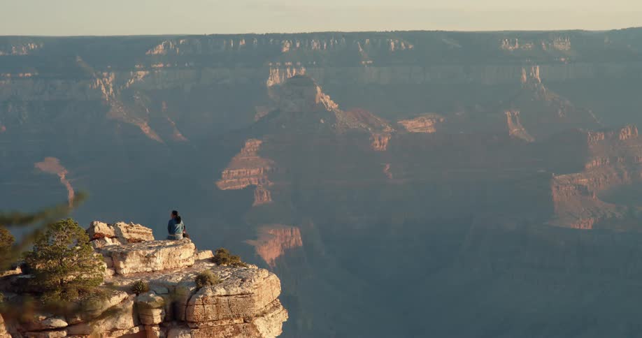 Someone sits on a ledge overlooking a mountain canyon and mountains and rock formations in the distance