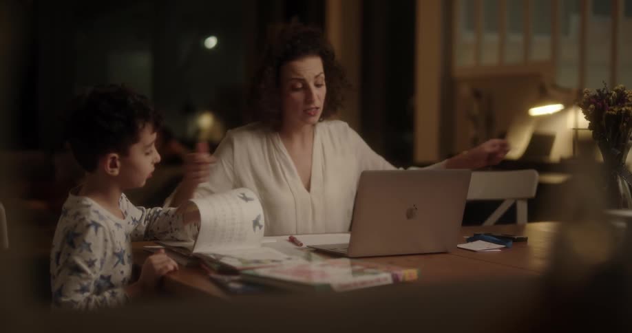 Mother and son sitting at the table while the son is studying and reading books and the mother is in a work related video call on her laptop