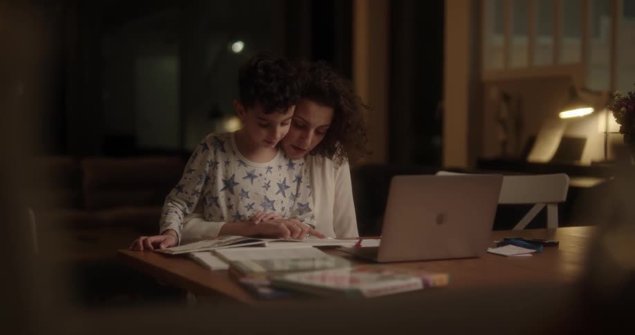 Mother helping her son read and study at the table with a laptop nearby