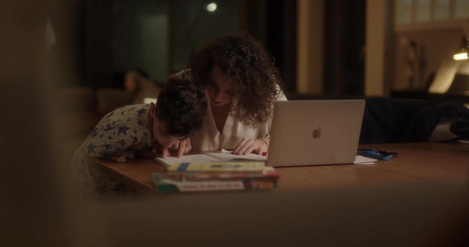 Mother sitting at table reads book with son next to laptop at night