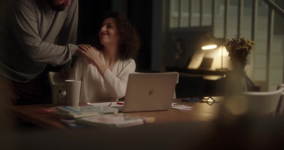 A man brings his wife a mug of coffee while she works late on her laptop