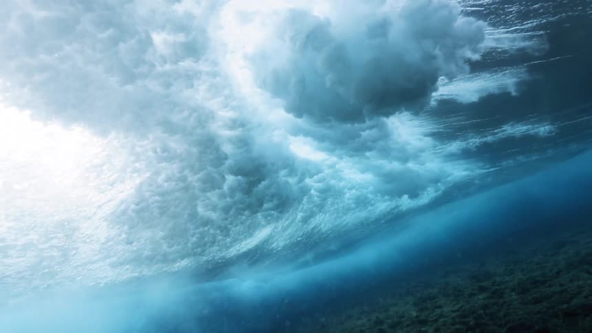 A rolling ocean wave is seen from below as it forcefully crashes down on the surface of the water