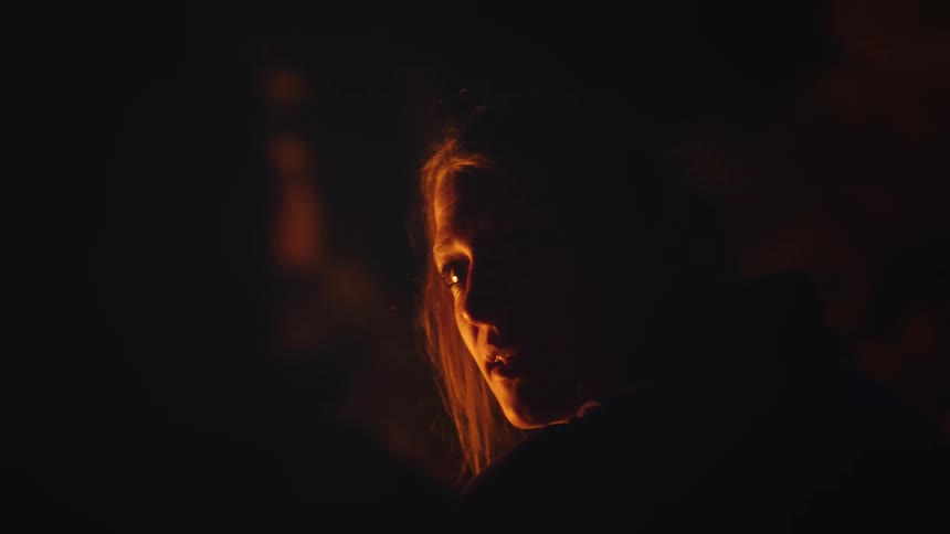 A woman sits in front of a fire at night and talks with someone next to her