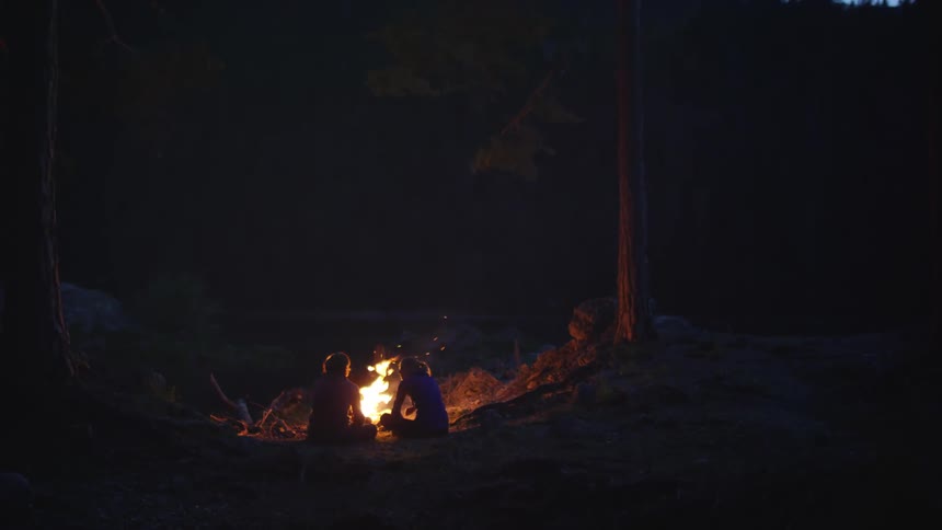 Woman and man sitting in the ground at a campfire near a forest