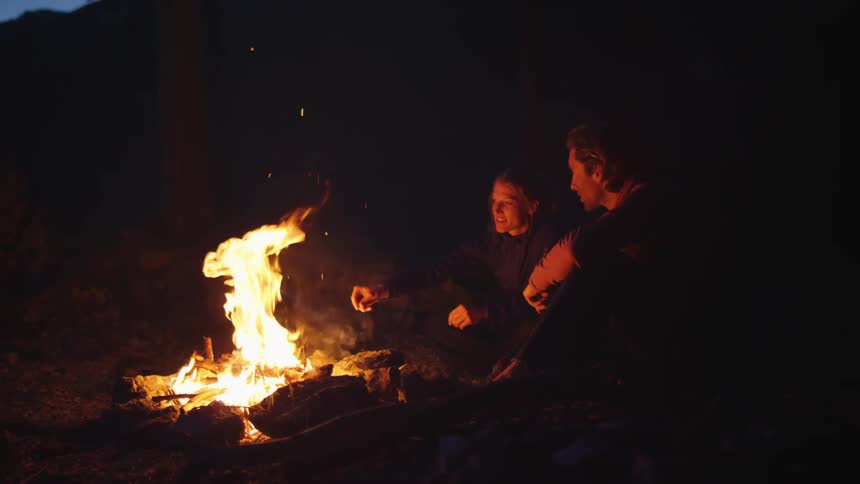 Man and woman sit on the ground next to a bonfire at night