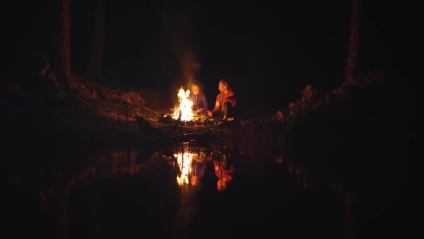 Two people are sitting next to a campfire along the side of a lake at night