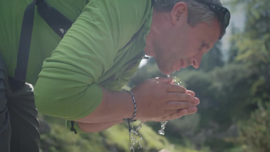 Man stands in a stream and splashes water on his face as two young girls crouch in the water nearby