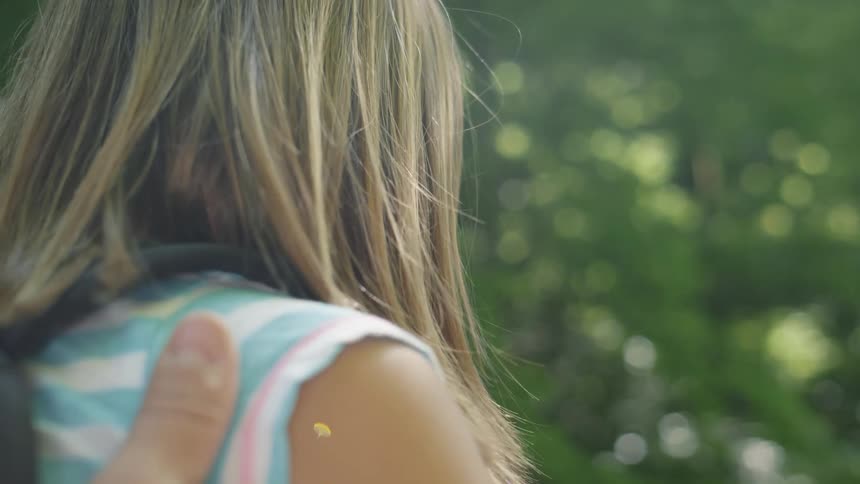 Young girl in a striped top is lifted by a hand while outdoors near the trees on a sunny day