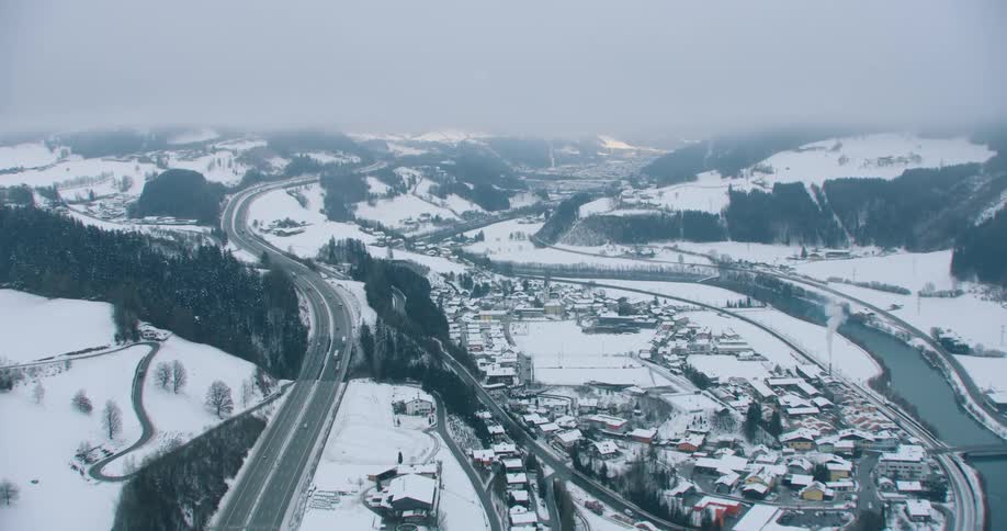 Vehicles drive down a highway that runs parallel to a river in a mountain town