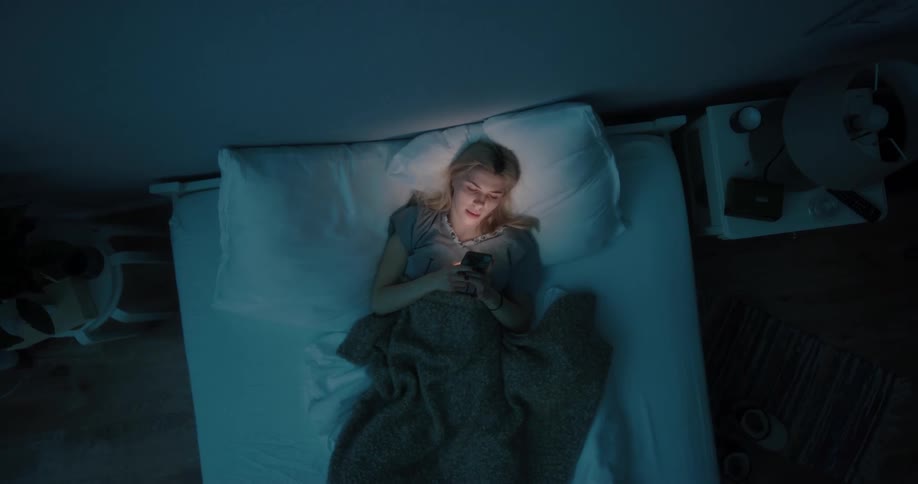Blond teenaged girl lays in bed and looks at her cell phone screen