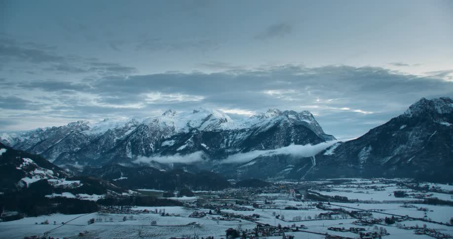 Traffic on a highway and snow covered mountains in the distance under a cloudy sky