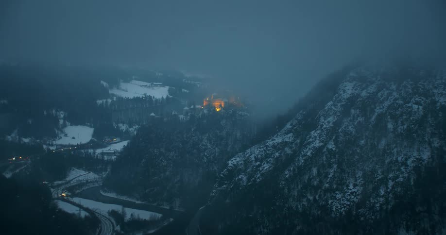 A building glows brightly on top of a ridge in a foggy snow covered valley